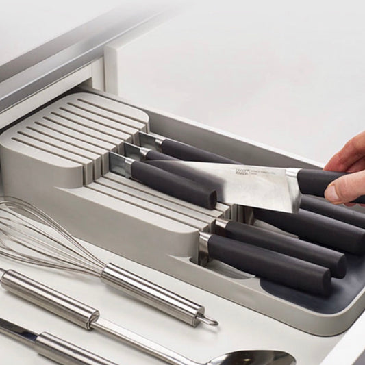 Eine Hand platziert ein Kochmesser in einen modernen Messer-Organizer in einer Küchenschublade. Der Messerhalter hat mehrere Schlitze unterschiedlicher Größe für verschiedene Messer, die parallel zueinander angeordnet sind. Neben dem Messer-Organizer liegen ein Schneebesen und ein Rührstab, die die Funktionalität und das durchdachte Design der Küchenutensilienaufbewahrung unterstreichen.