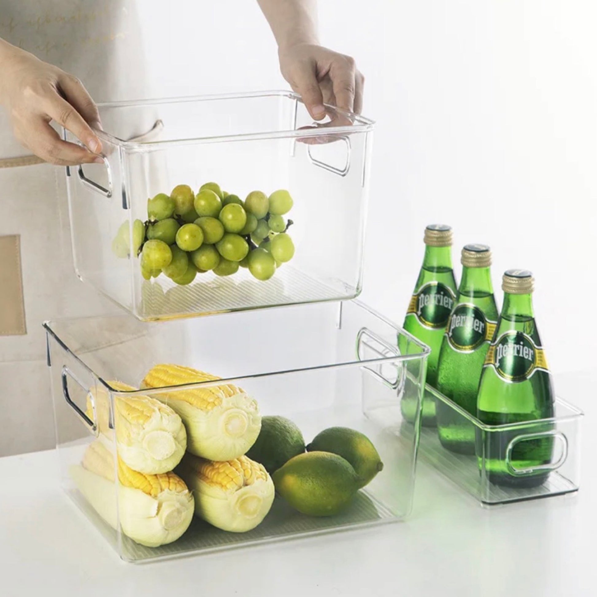 Hände heben eine transparente Aufbewahrungsbox aus einem Regal, gefüllt mit grünen Trauben. Darunter sind zwei weitere Boxen: eine mit frischen Maiskolben und Limetten, die andere mit grünen Glasflaschen von Perrier. Diese Boxen sorgen für eine ästhetische und funktionale Art der Lebensmittellagerung in einem modernen Haushalt.