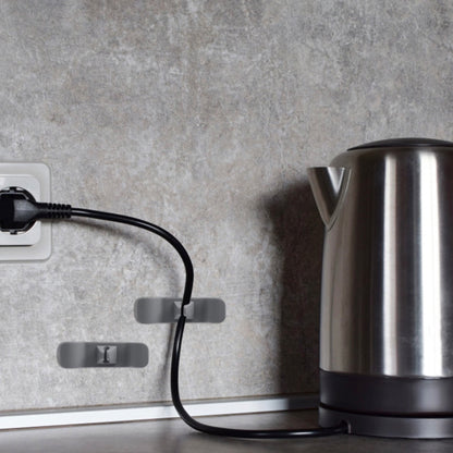 Das Bild zeigt eine Küchenszene mit einem Edelstahl-Wasserkocher auf einer Arbeitsplatte und zwei grauen Kabelhaltern an der Wand, die das Kabel des Wasserkochers ordentlich führen. Die Kabelhalter sind strategisch platziert, um das Kabel sauber und aus dem Weg zu halten, während der Wasserkocher in Gebrauch ist.