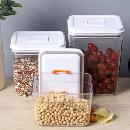 Transparente Vorratsdosen mit weißem Deckel und innovativem Silikonbandverschluss in Orange, der das Löffelhaltesystem darstellt. Eine Dose ist geöffnet und zeigt trockene Kichererbsen im Vordergrund, während im Hintergrund andere Dosen mit verschiedenen Trockenfrüchten und Körnern die vielseitige Nutzung für eine ordentliche und ansprechende Lebensmittelaufbewahrung demonstrieren.