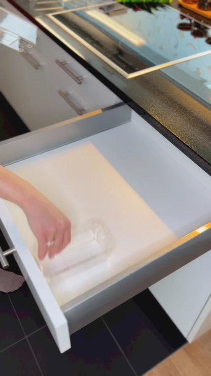 Video zeigt eine Person, die einen transparenten Schubladen-Organizer in einer Küchenschublade einrichtet. Zuerst werden die einzelnen Teile des Organizers in die Schublade eingesetzt. Anschließend füllt die Person die Fächer sorgfältig mit verschiedenen Küchenartikeln, darunter Teebeutel, Kaffeekapseln, Servietten, Lindt Bonbons und Kakaopulverbeutel. Das Video demonstriert eine effiziente Methode zur Organisation und leichten Zugänglichkeit dieser Artikel in der Küche.