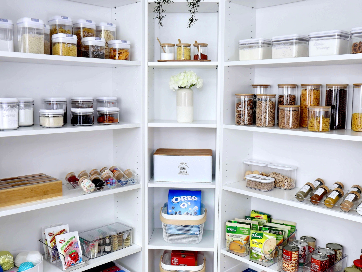 Elegante weise Speisekammer, gefüllt mit verschiedenen Vorratsdosen und Aufbewahrungsboxen für Lebensmittel wie Pasta, Getreide, Snacks und Konserven – perfekt organisiert für mehr Übersicht und Ordnung in der Küche.