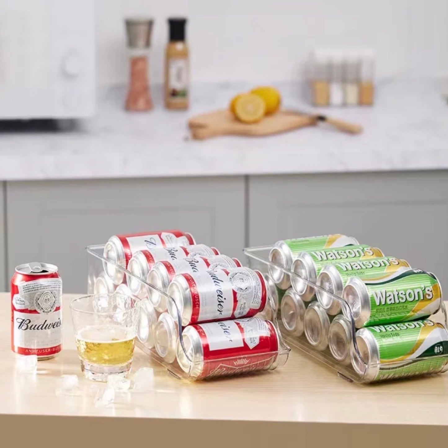 Zwei transparente Dosenhalter auf einer Küchenarbeitsplatte, gefüllt mit roten Budweiser-Bierdosen und grünen Watson’s-Sodadosen, neben einem halb leeren Bierglas und einer umgestürzten Dose, im Hintergrund Gewürze und eine Zitrone auf einem Schneidebrett.