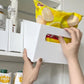 Hände entnehmen eine weiße Aufbewahrungsbox mit Griff, gefüllt mit Lebensmittelpackungen, aus einem Küchenschrank.
