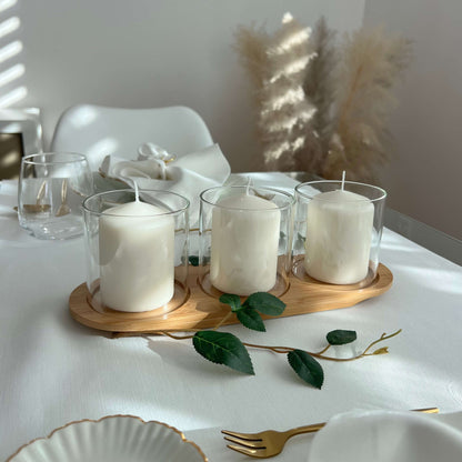 Drei Windlichtgläser mit weißen Kerzen auf einem ovalen Holztablett, dekoriert mit grünem Zweig auf einem gedeckten Tisch mit goldfarbenem Besteck und stilvollen Details.