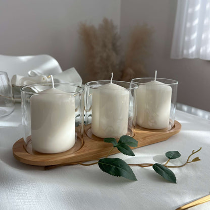 Drei Windlichtgläser mit weißen Kerzen auf einem ovalen Holztablett, dekoriert mit einem grünen Zweig auf einer festlich gedeckten Tafel.