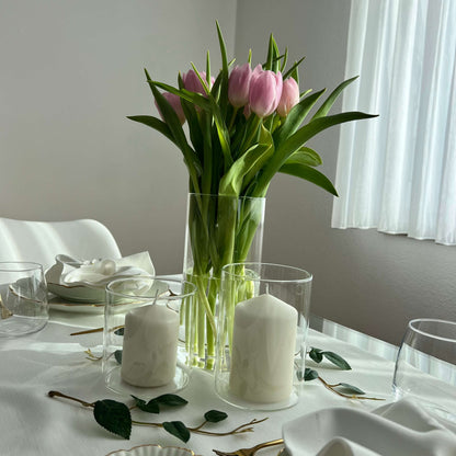 Zwei Windlichter mit weißen Kerzen und eine hohe Vase mit rosa Tulpen auf einem elegant gedeckten Tisch mit weißer Tischdecke und Servietten.