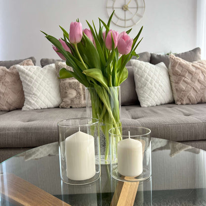 Zwei Windlichter mit weißen Kerzen und eine Glasvase mit rosa Tulpen auf einem runden Couchtisch vor einem Sofa mit Kissen in neutralen Tönen