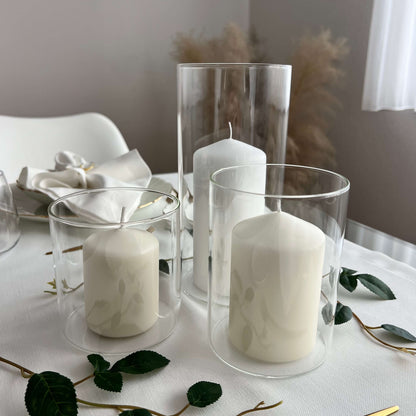 Drei Windlichter aus Glas in unterschiedlichen Größen mit weißen Kerzen, dekorativ arrangiert auf einem weiß gedeckten Tisch mit grünen Blättern als Akzent.