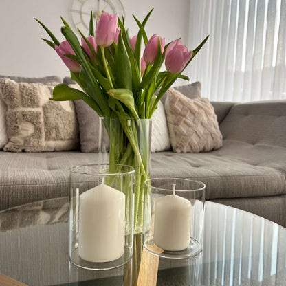 Zwei Windlichter mit weißen Kerzen und eine Glasvase mit rosa Tulpen auf einem runden Couchtisch vor einem Sofa mit Kissen in neutralen Tönen.