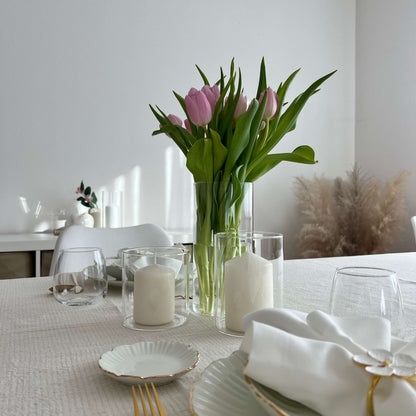 Windlichter mit weißen Kerzen und hohe Glasvase mit rosa Tulpen auf einem festlich gedeckten Tisch mit weißem Geschirr und goldfarbenem Besteck.