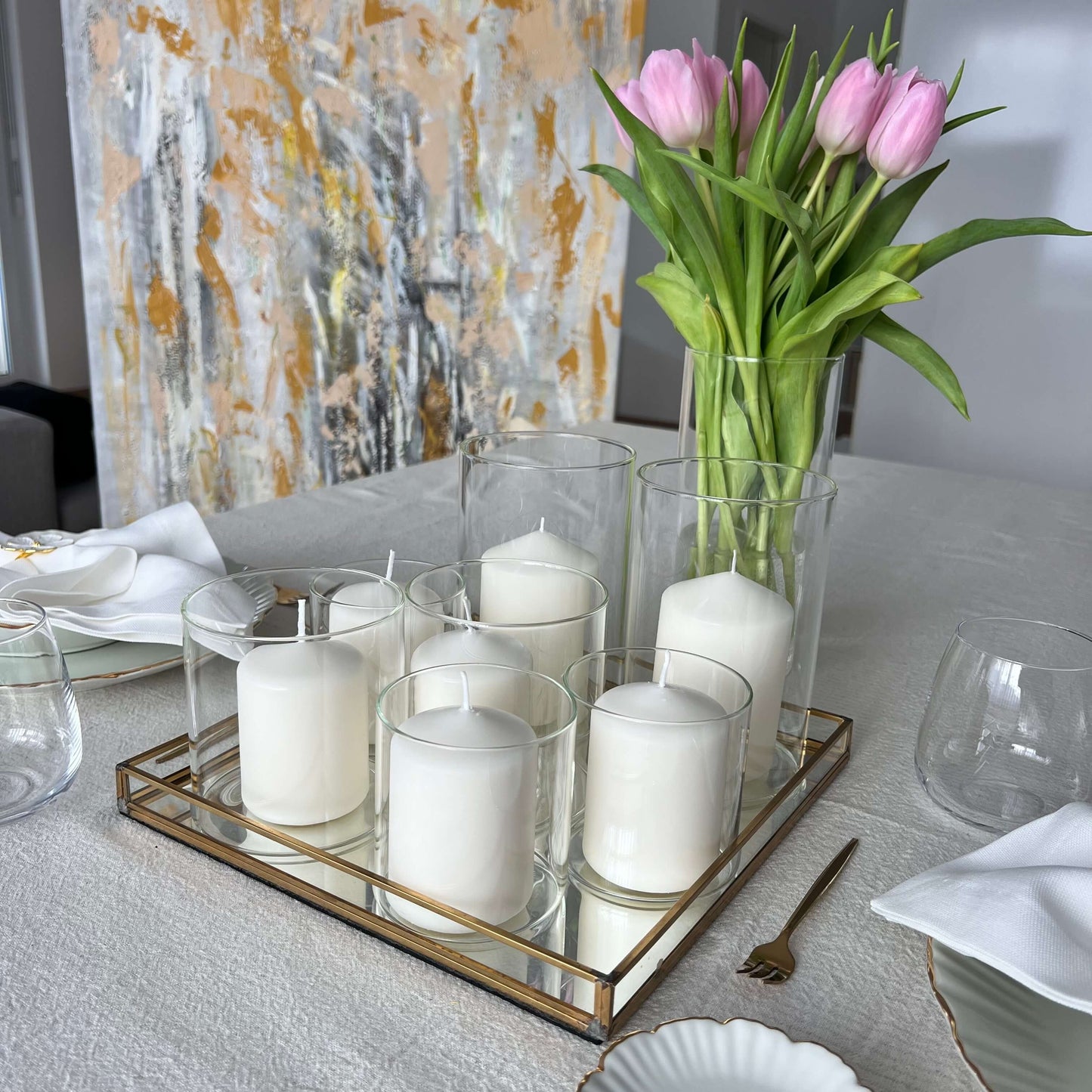 Windlichter aus Glas mit weißen Kerzen auf einem goldgerahmten Tablett, dekoriert mit rosa Tulpen in einer Vase und einem abstrakten Wandbild im Hintergrund